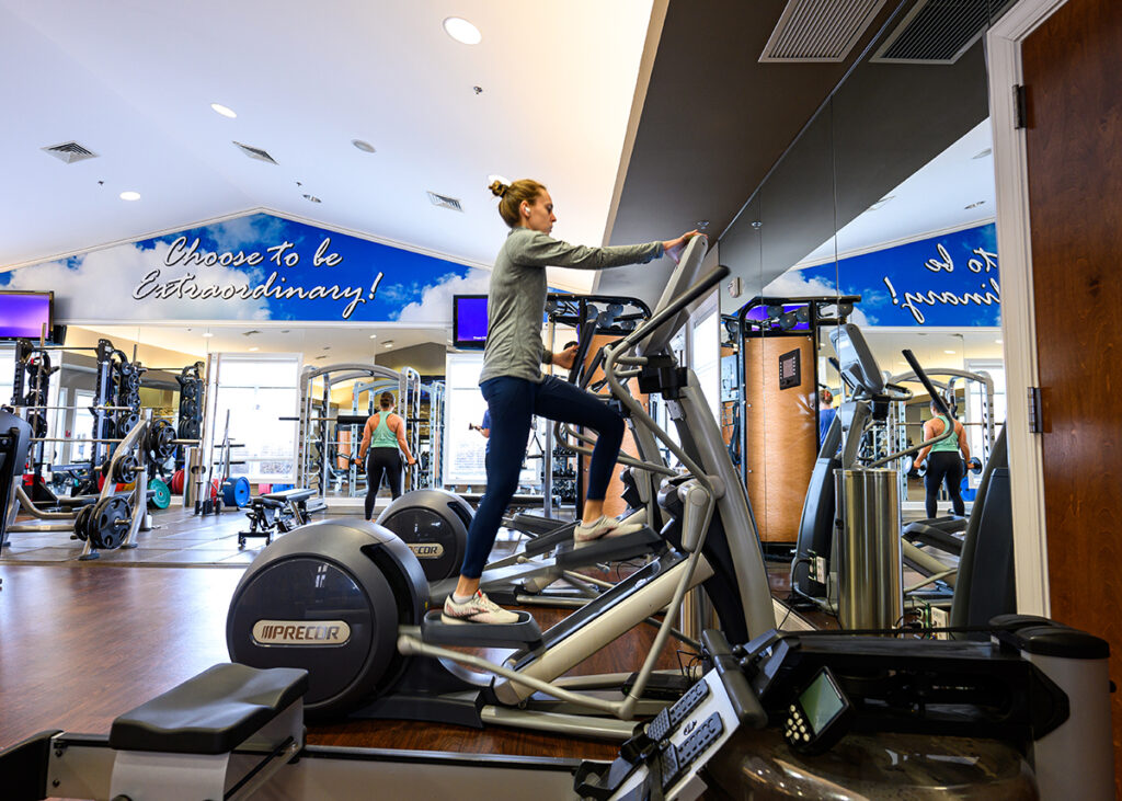 Pictured is Polett Rosztoczy, assessment and training coordinator, exercising on an elliptical trainer in the HPU Employee Wellness Center. In the background is Austen Smith, director of campus fitness for Campus Recreation, doing biceps curls.