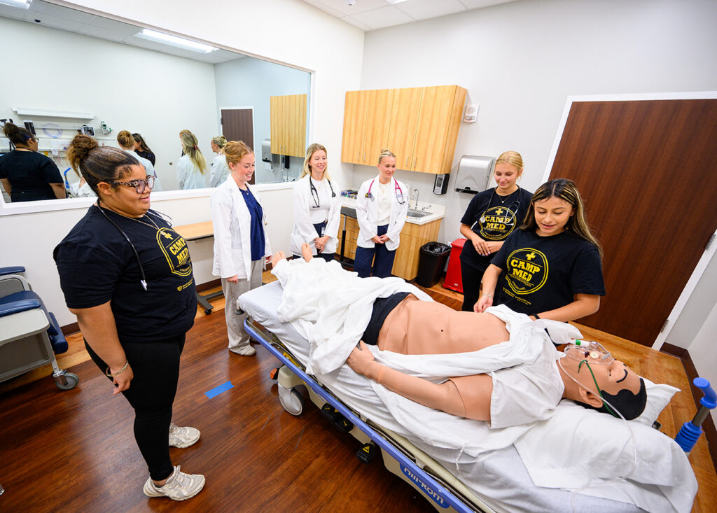 Shown at center in white coats, HPU PA students McKenna Roof, Ana Haselden and Alyssa Avallone smiled as high school students experienced a patient simulation lab during CampMed.